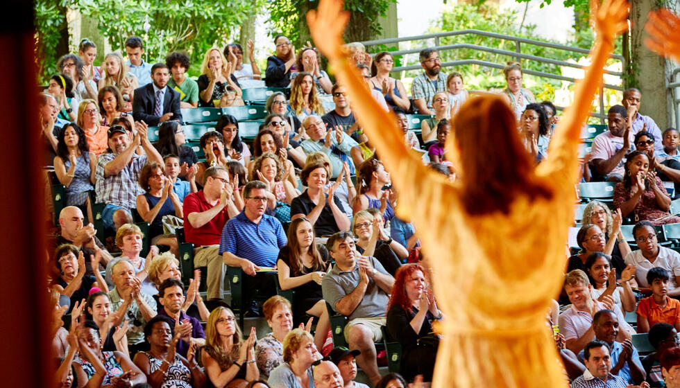 The back of a young performer on stage. She is facing the audience with arms raised