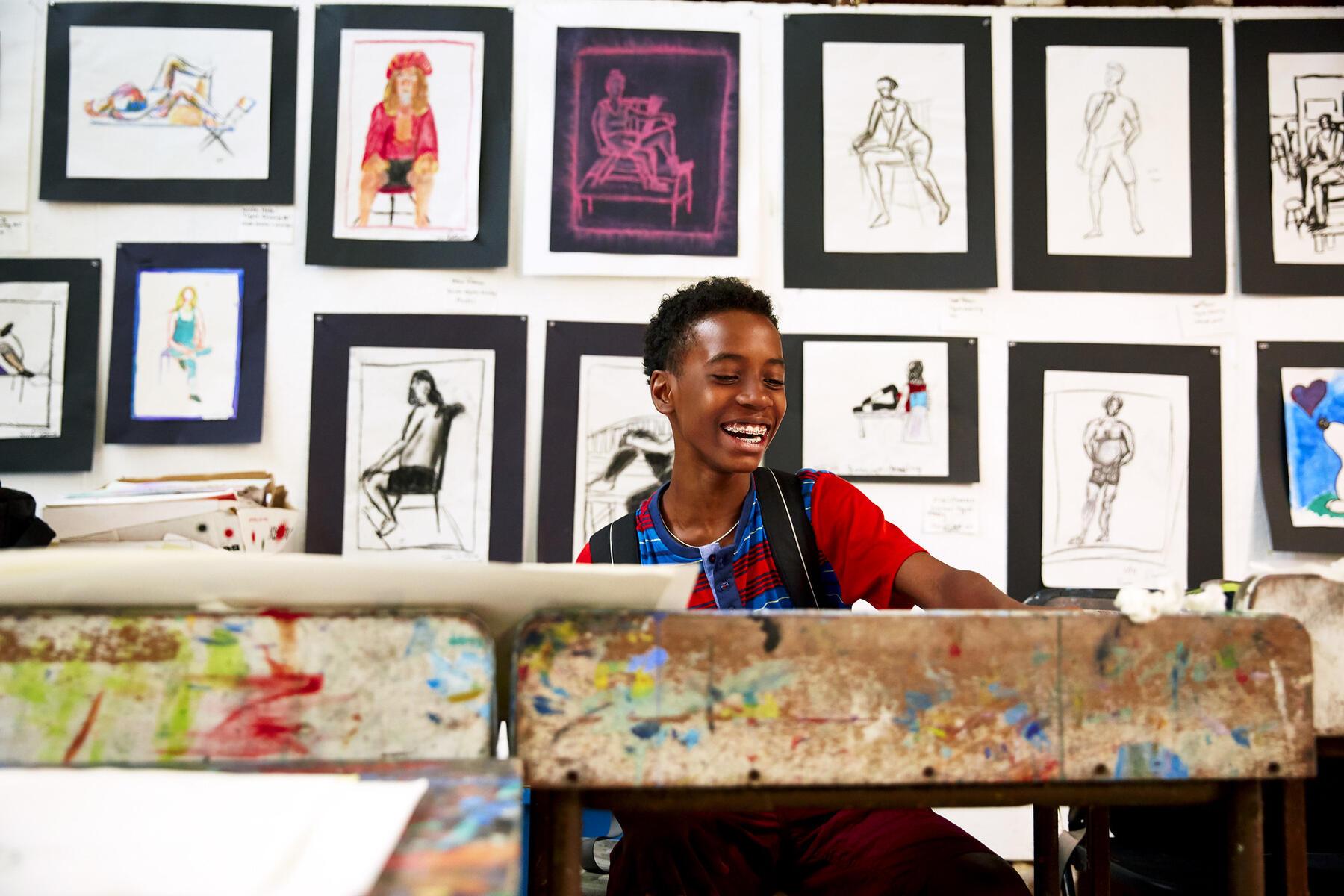 Young boy sitting at an art studio desk smiling with paintings pinned up behind him
