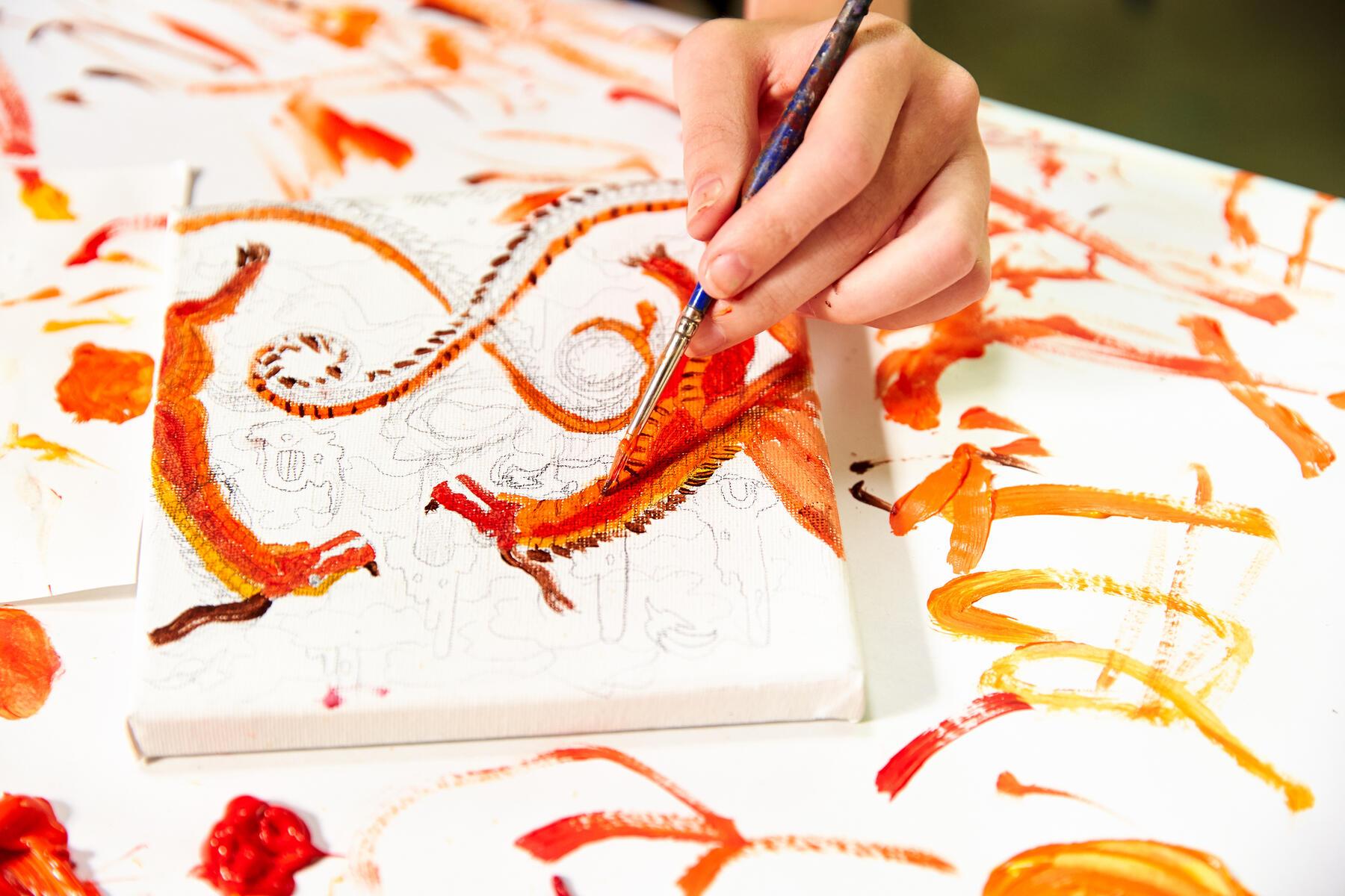 Closeup of child's hand painting an orange dragon on canvas