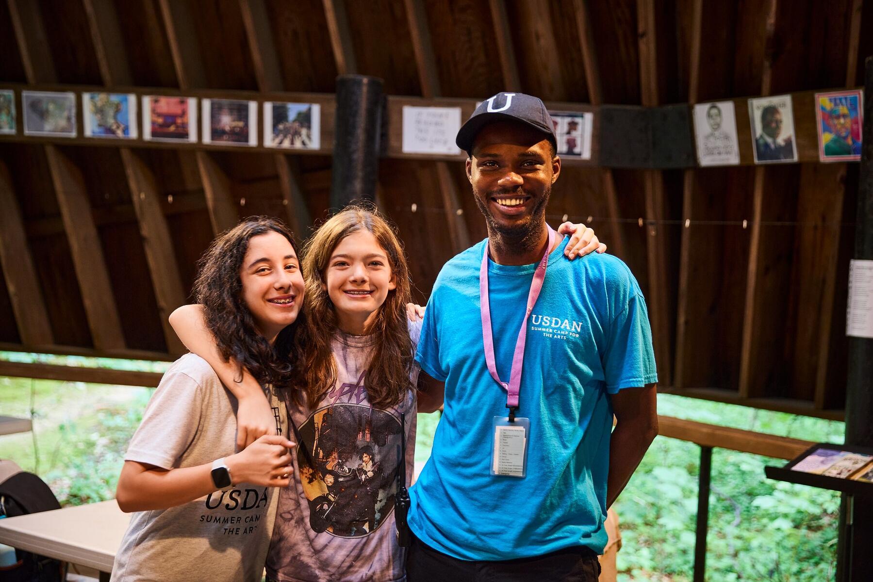Two female-presenting students with their arms around each other standing with a teacher in a blue Usdan shirt