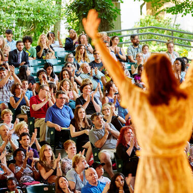 The back of a young performer on stage. She is facing the audience with arms raised